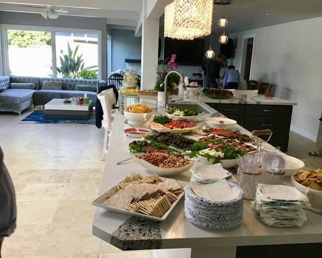 A buffet table with a variety of food items including salads, crackers, and meats is set up in a modern kitchen, with a spacious living area visible in the background.