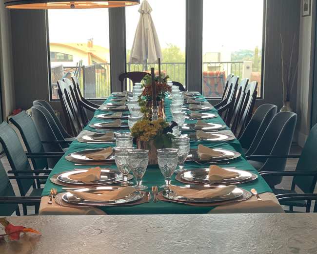 A long dining table is set with plates, glasses, and napkins, surrounded by chairs, under a large window with a view outside.