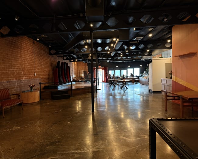 The image shows an industrial-style interior space with exposed brick walls, polished concrete floors, and various seating arrangements along with a wooden stage area and tables set up for gathering.