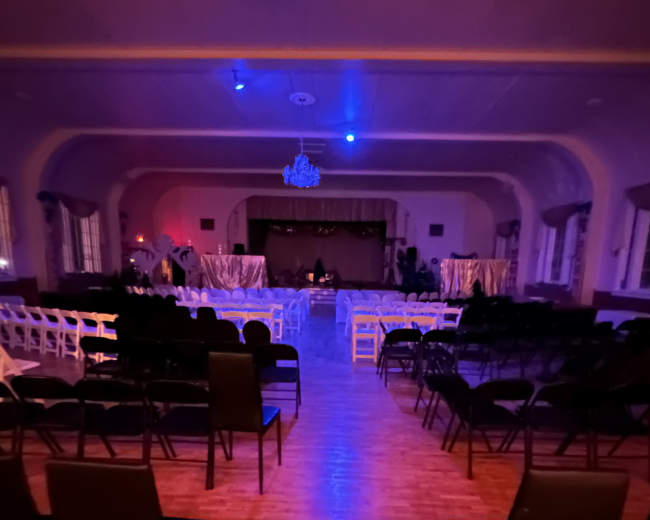 A dimly lit auditorium setup with rows of empty chairs facing a stage, illuminated by blue lights.