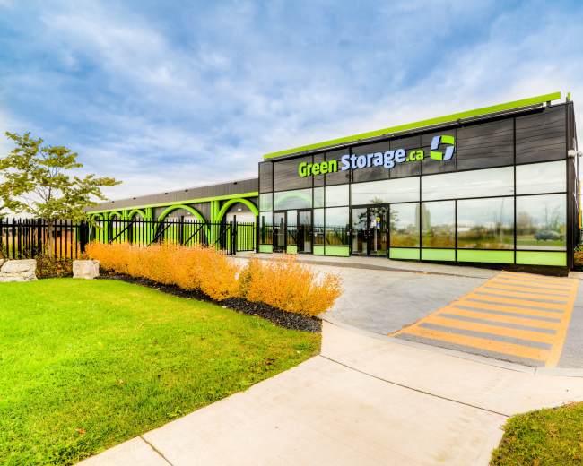 The image shows a modern storage facility with large glass windows and a green and black exterior, situated next to a landscaped area.