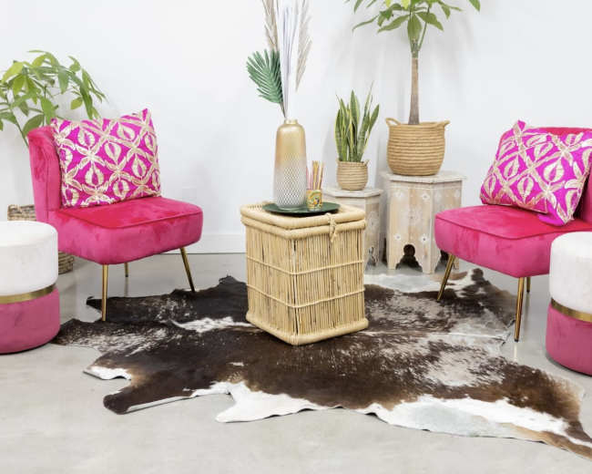 The image depicts two pink chairs with patterned cushions flanking a wicker coffee table, surrounded by plants and a cowhide rug in a bright, minimalist room.