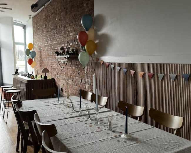A long dining table set with candles and a checkered tablecloth is positioned against a brick wall, decorated with balloons and a bunting banner.