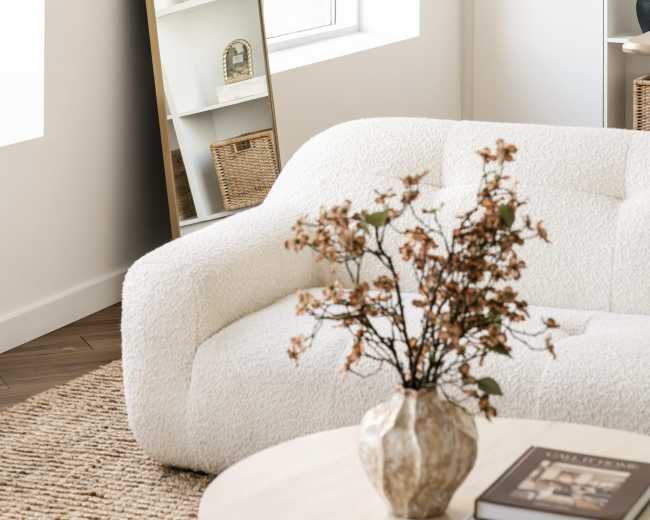 A modern living room features a plush white sofa, a round coffee table, and a tall mirror leaning against a wall, with a decorative vase of dried flowers on the table.