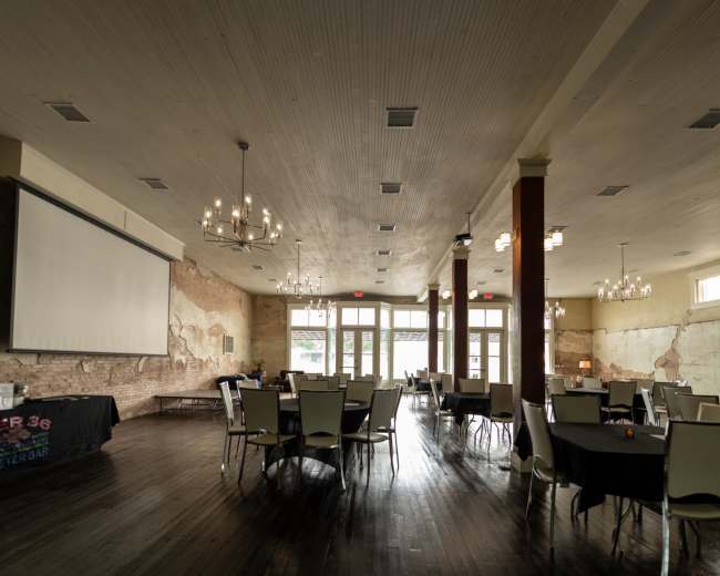 The image shows a spacious indoor event venue with tables set for dining and a large projector screen mounted on a wall.