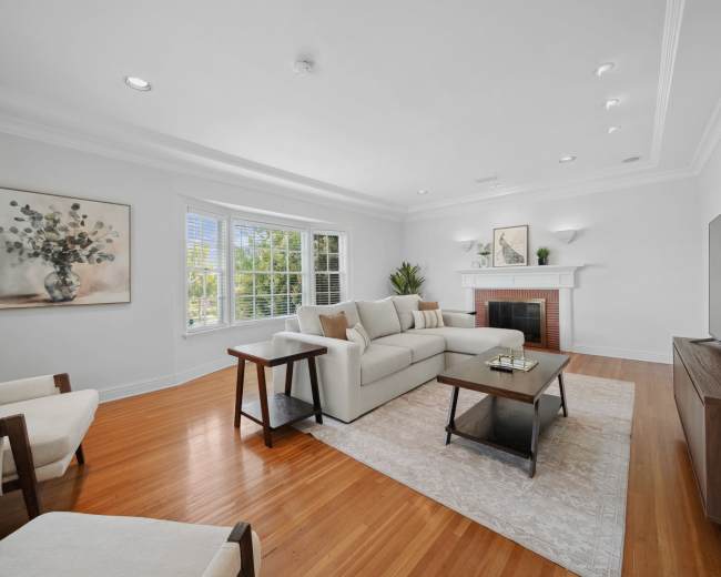 A spacious living room features a light-colored sofa, a coffee table, and a television, with large windows bringing in natural light.
