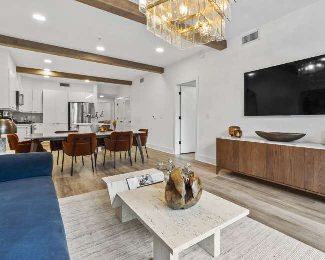A modern living room features a blue sofa, a wooden coffee table, a large television mounted on the wall, and a dining area with a table and chairs in the background.