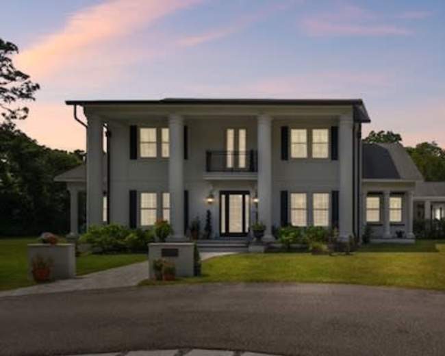 The image depicts a modern two-story house with a large front porch, flanked by a single-story wing, set on a landscaped lawn near a circular driveway.