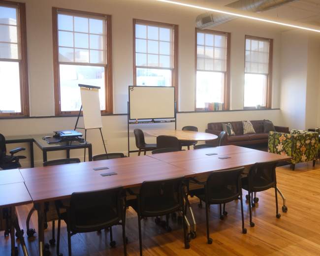The image shows a modern office space with large windows, a circular table, a sofa covered in floral fabric, and several chairs arranged around adjustable desks.