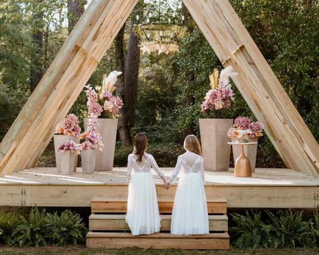 Two girls in white dresses stand hand-in-hand on wooden steps in front of a triangular wooden structure adorned with floral arrangements.