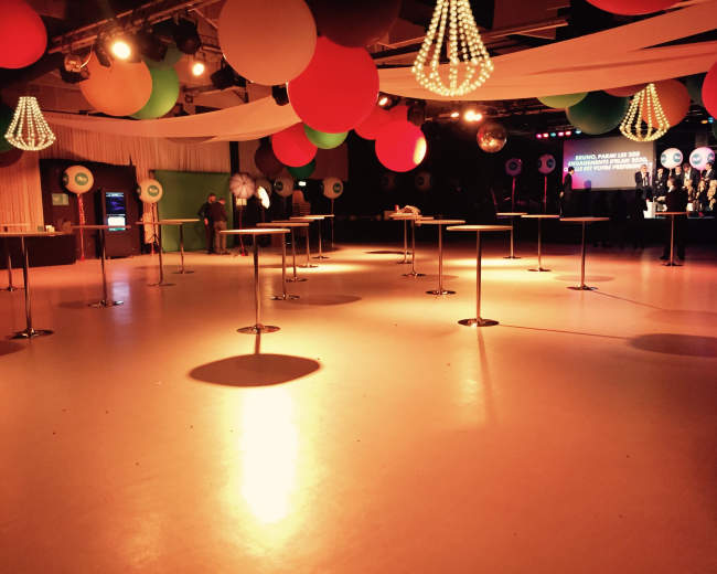 The image shows an empty event space with round tables, colorful hanging decorations, and a stage at the back.