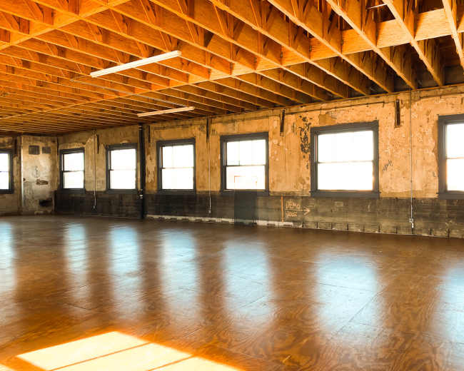 The image shows a spacious, empty room with wooden floors and large windows, characterized by exposed beams and a weathered interior.