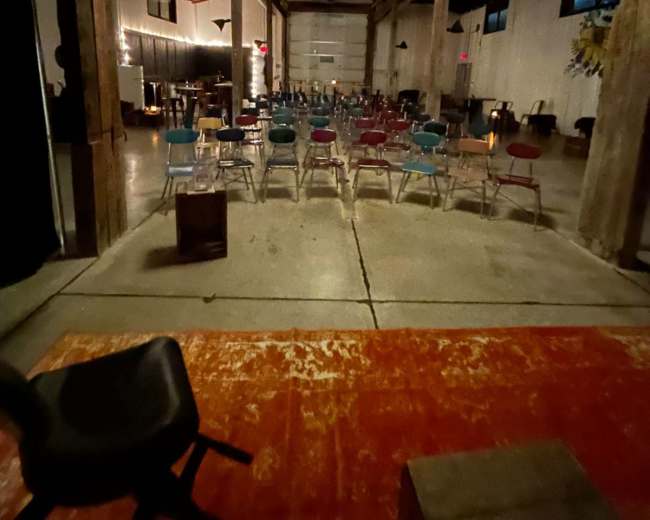 The image shows a spacious indoor venue with rows of colorful chairs arranged facing a stage, illuminated by hanging chandeliers and featuring exposed wooden beams and a concrete floor.