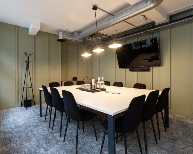 A modern conference room features a long white table surrounded by black chairs, with a large screen mounted on the wall and warm overhead lights.