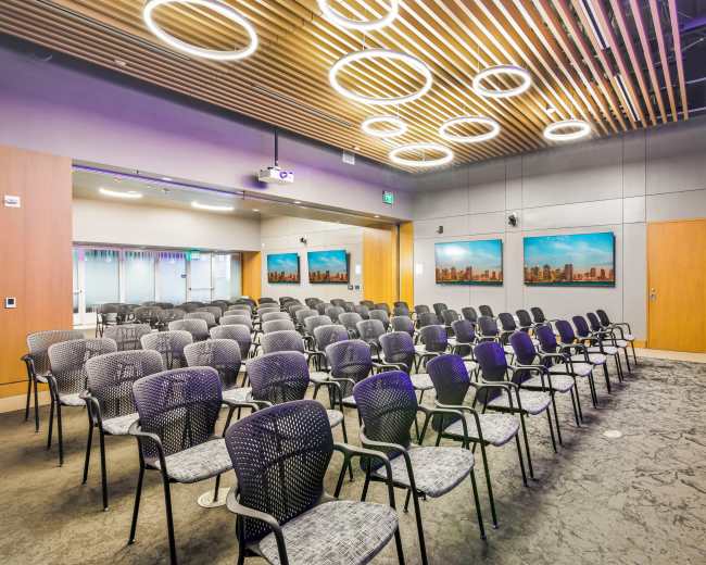 A modern conference room is arranged with rows of black chairs facing a projection screen and features art on the walls.