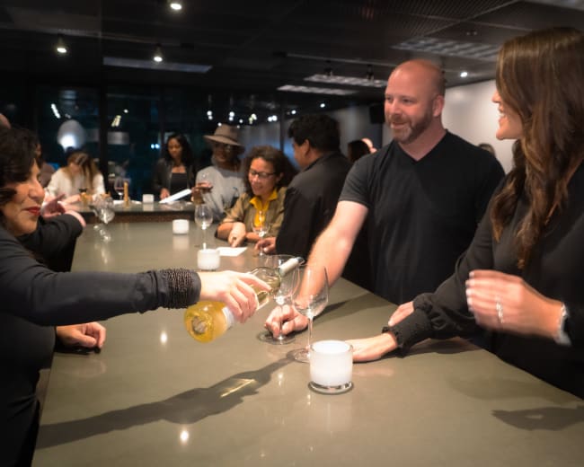 A group of people gathers around a bar, engaging in conversation and enjoying drinks while one person is pouring a beverage into a glass.