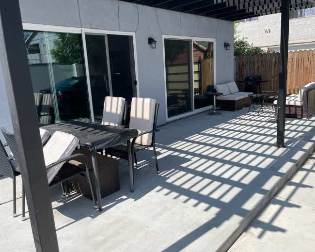 A patio area with a table and chairs, sofas, and large sliding glass doors is shown under a partially shaded pergola.