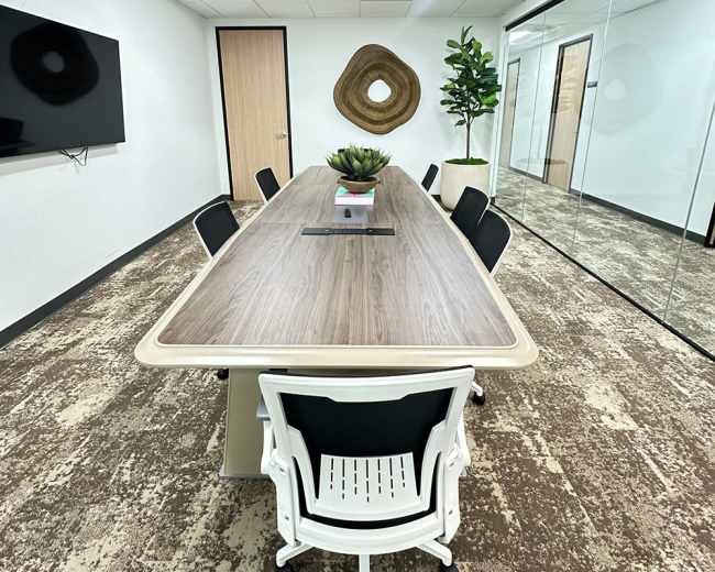 The image shows a modern conference room with a long table surrounded by white chairs, a wall-mounted TV, and a large glass panel separating it from an adjacent area featuring a decorative plant.