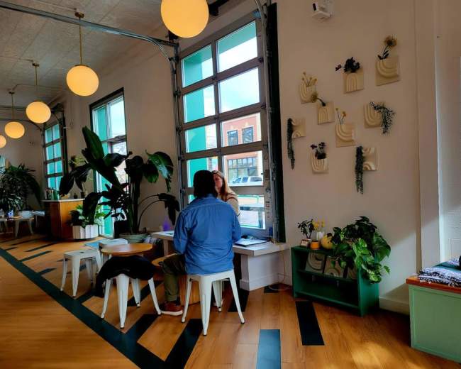 A person sits at a table in a bright, modern café, engaged in conversation with another person across from them, surrounded by large windows and decorative plants.