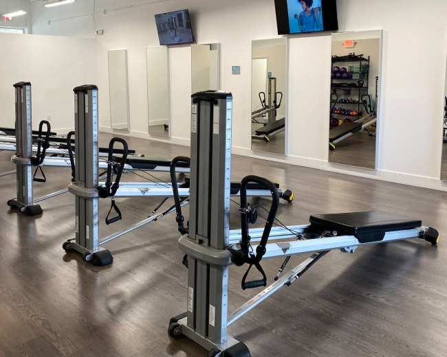 The image shows a modern fitness studio with several rowing machines aligned in a well-lit space featuring mirrors and wall-mounted screens.