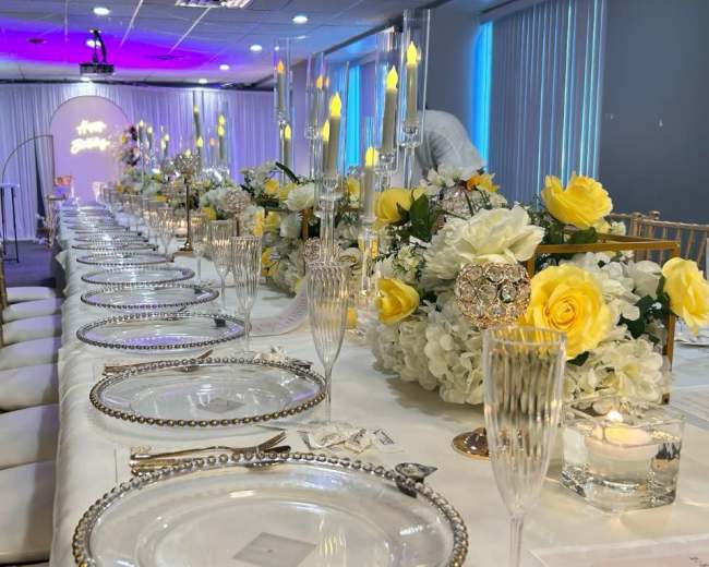 A long banquet table is elegantly arranged with decorative plates, crystal glassware, and floral centerpieces featuring yellow roses and white flowers.