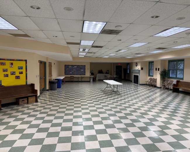 The image shows a large, empty room with a checkered floor, foldable tables, and a few benches along the walls.
