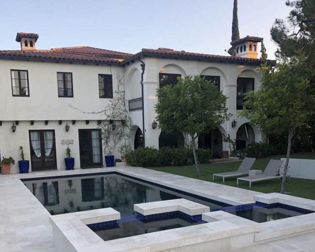 The image shows a modern home with a two-story facade overlooking a rectangular pool surrounded by a stone patio and landscaped yard.