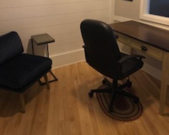 A small, minimalist office space with a wooden desk and a black office chair, accompanied by a blue-striped armchair and a small side table.