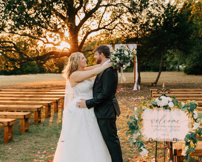 A couple stands together in an outdoor wedding venue adorned with wooden benches and a floral welcome sign, with the sun setting in the background.