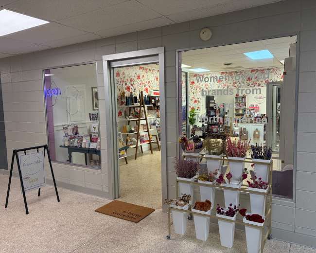 The image shows a retail store display with floral wallpaper and products arranged on shelves, featuring a sign that highlights women-owned brands from North America.
