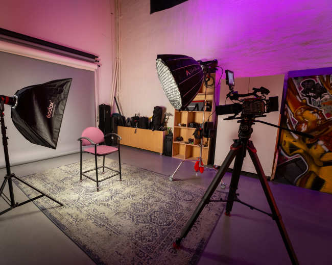 The image shows a photography studio setup with a central chair, softbox lights, a camera on a tripod, and equipment displayed in the background.