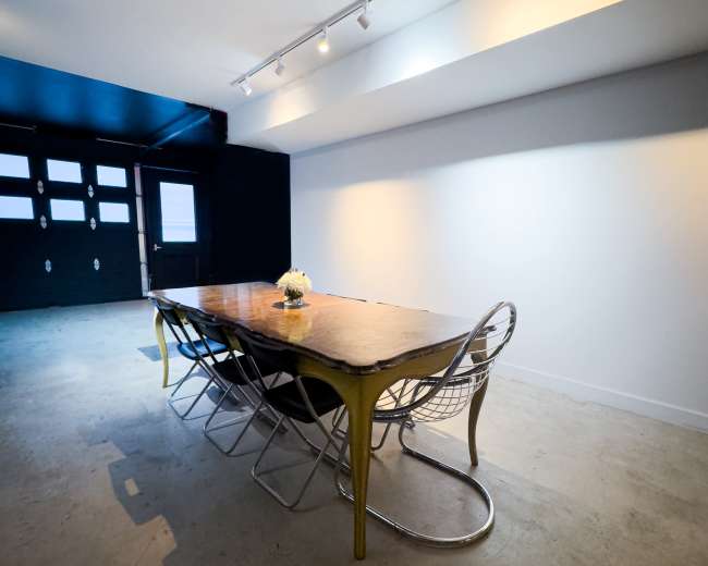 A long table with metal chairs is placed in a spacious room featuring a gray floor and a dark blue ceiling, illuminated by track lighting.