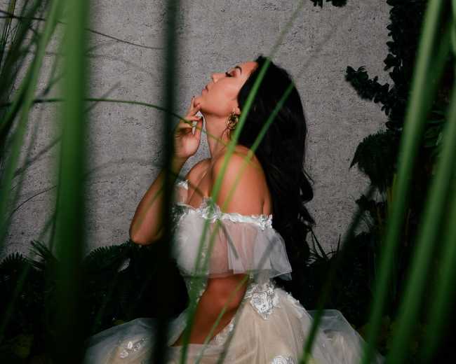 A woman in a white off-the-shoulder dress, seated among tall greenery against a textured wall.