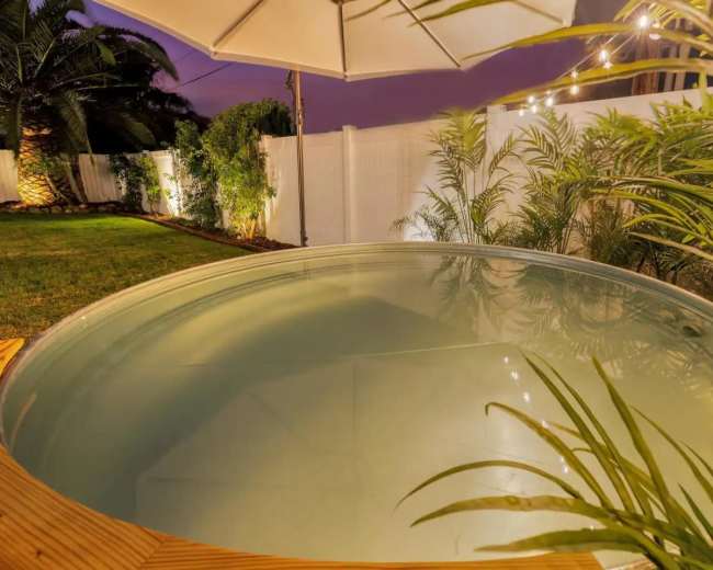 A round hot tub is set within a backyard surrounded by lush greenery and a white fence, illuminated by soft outdoor lighting.