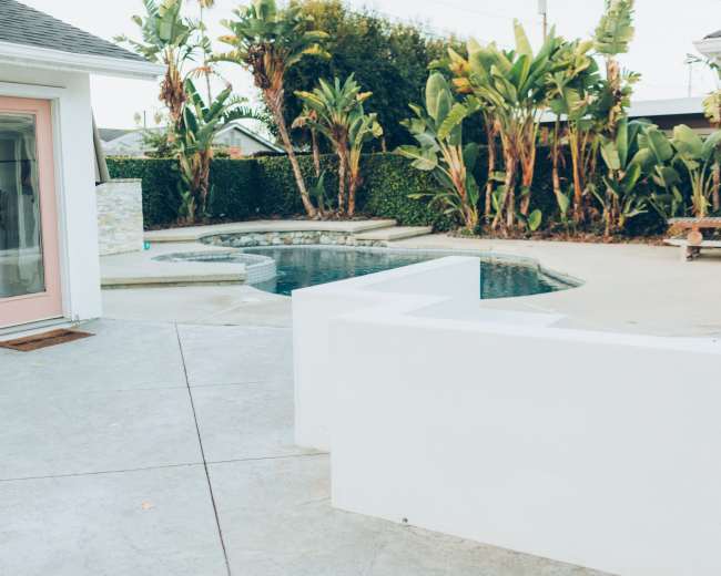 The image shows a backyard with a pool surrounded by palm trees and a white structure with a pink door.