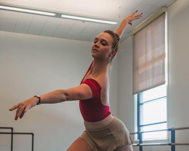A ballet dancer performs an expressive pose in a studio with a wooden floor and ballet barres in the background.