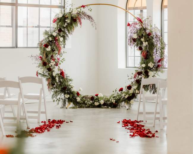 A circular floral arrangement adorned with red and white flowers stands as a backdrop in a bright, minimalist wedding venue with white chairs arranged in front of it.