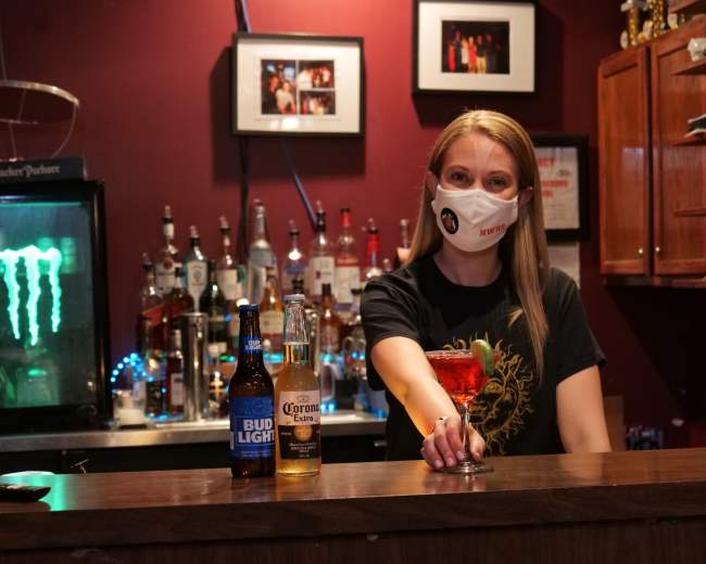 A bartender wearing a mask stands behind a bar, holding a cocktail while two beer bottles are placed on the counter.