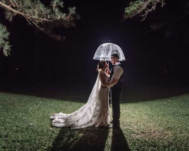 A bride and groom hold a clear umbrella while standing on a grassy area at night, illuminated by a light behind them.