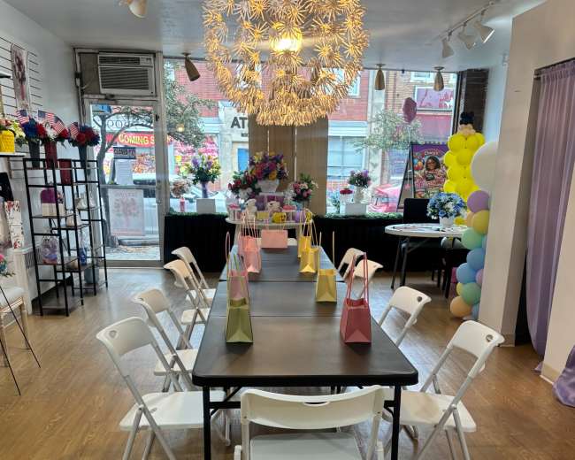 A spacious indoor event area features a long black table surrounded by white folding chairs, decorated with colorful gift bags and a large decorative chandelier.
