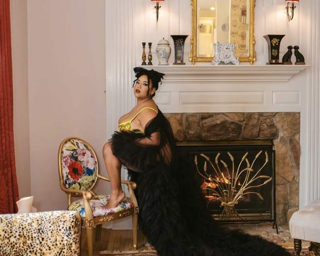 A woman wearing an elaborate gown poses in front of a fireplace, seated on a decorative chair in a stylishly furnished room.