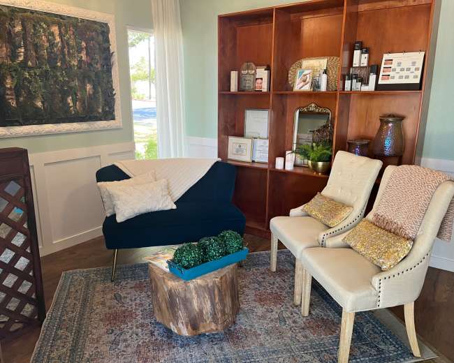 A cozy interior with a black couch and two cream-colored chairs arranged around a wooden stump table on a decorative rug, accompanied by a bookcase and a wall adorned with greenery.