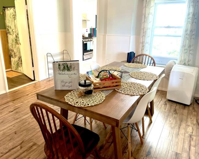 A dining table with placemats and a welcome sign is set in a well-lit room with a kitchen doorway and a fan visible in the background.