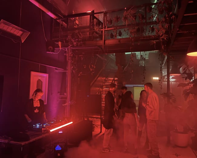 A DJ performs in a dimly lit venue filled with colorful lights and smoke while a small group of people dance nearby.