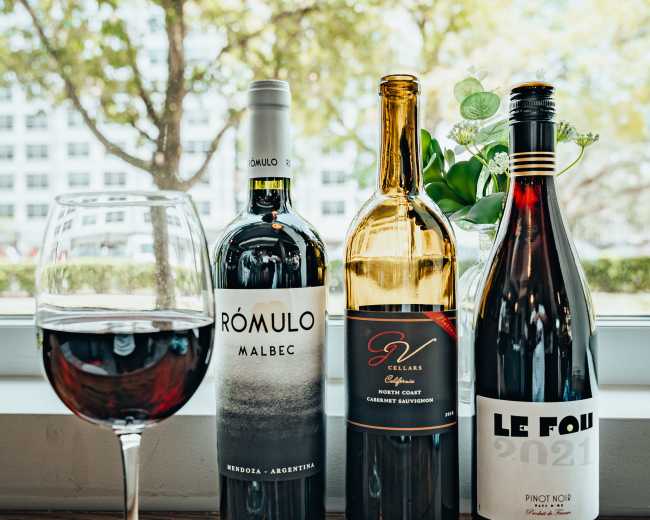 The image shows three bottles of wine — a Malbec, a Cabernet Sauvignon, and a Pinot Noir — placed on a wooden surface beside a filled wine glass, with trees visible through a window in the background.