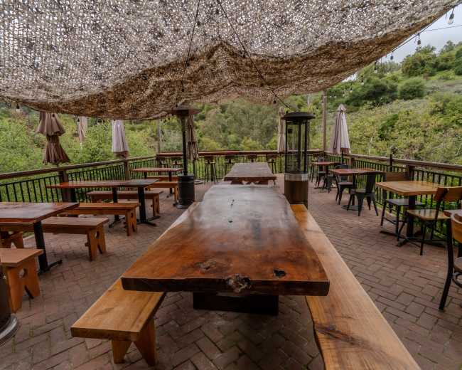 The image shows a wooden outdoor dining area with a large table, benches, and shaded by a canopy, surrounded by greenery.