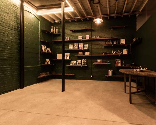 The image shows a room with green brick walls, wooden shelves displaying various items, and a round wooden table positioned against the wall.