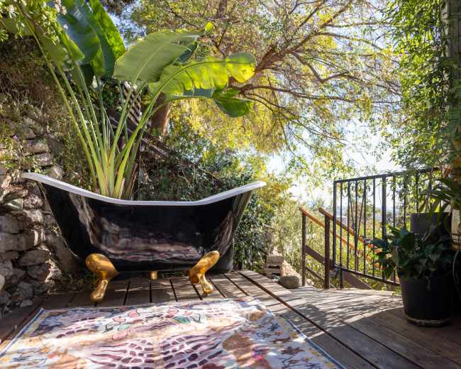 A vintage clawfoot bathtub filled with large tropical plants sits on a wooden deck surrounded by greenery and colorful foliage.