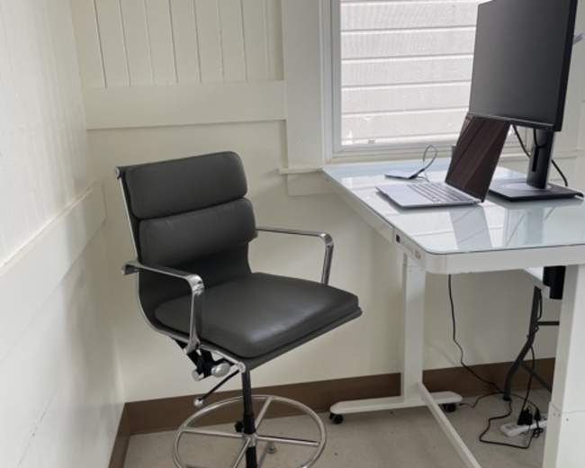 A black bar stool is positioned next to a desk with a computer monitor in a bright room.