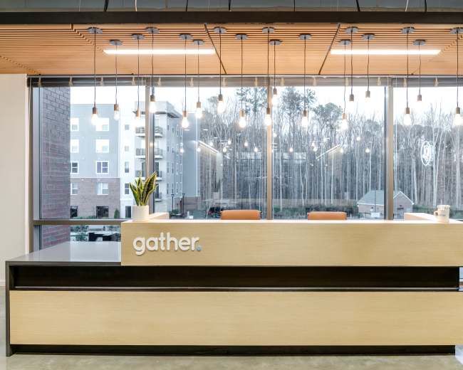 A modern reception desk with the word "gather" prominently displayed sits in front of large windows overlooking a wooded area.